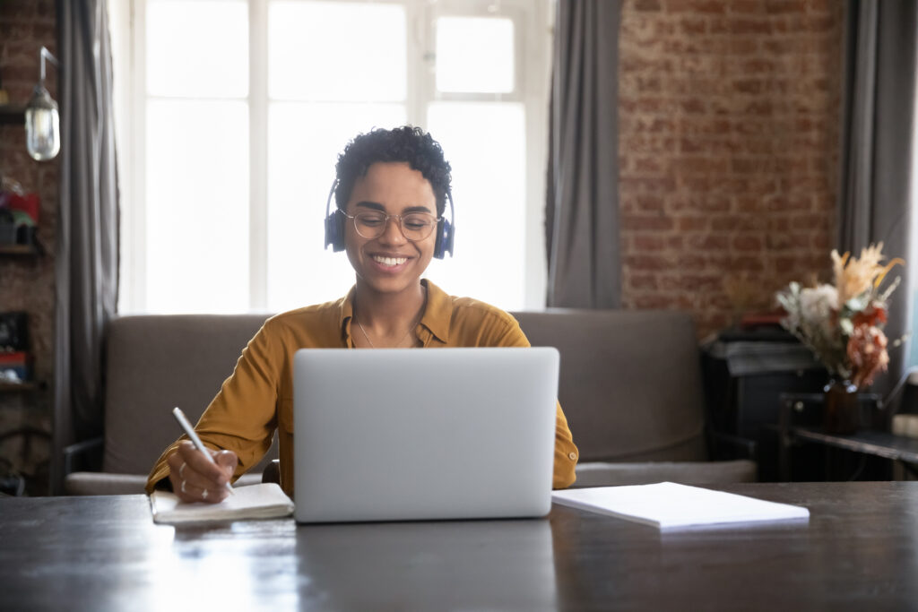 Happy interested young African American woman in glasses and headphones watching educational webinar or online lecture, remembering information writing notes in copybook, preparing for exams at home.